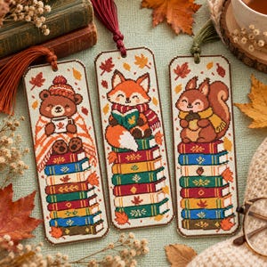 May include: Three rectangular cross-stitch bookmarks featuring embroidered animals. The left shows a bear with a mug, the center a fox reading a book, and the right a squirrel holding an acorn. Each sits atop a stack of colorful books with autumn leaves.