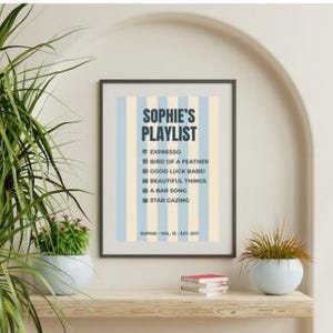 May include: A framed rectangular wall art print featuring vertical light blue and cream stripes. The text reads SOPHIE'S PLAYLIST followed by a list of song titles including EXPRESSO, BIRD OF A FEATHER, GOOD LUCK BABE, BEAUTIFUL THINGS, A BAR SONG, and STAR GAZING.