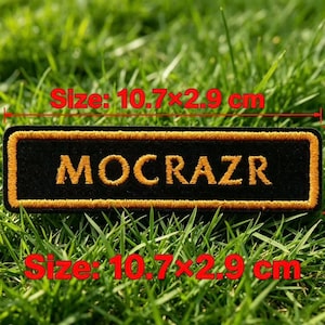 May include: A rectangular black embroidered patch featuring the text MOCRAZR in gold thread, bordered by a gold stitched frame. The patch measures 10.7 by 2.9 cm as indicated by the red text and lines overlaid on the image.