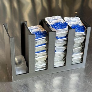 May include: A black 3D-printed plastic organizer with four vertical slots designed to hold stacks of contact lens blister packs. The organizer contains multiple Acuvue Oasys for Astigmatism contact lens packages with blue and white labels.