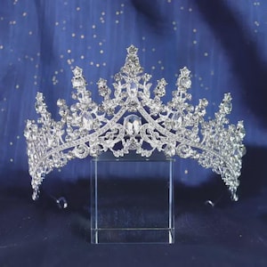 Könnte beinhalten: Ein silberfarbenes Metall-Diadem mit einem komplizierten, symmetrischen Design, das mit zahlreichen klaren, facettierten Kristallen verziert ist. Die Krone hat eine hohe Mittelspitze und mehrere kleinere Spitzen, die mit funkelnden Steinen besetzt sind.