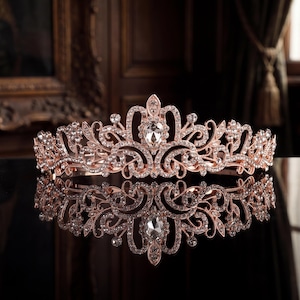 May include: A rose gold tiara featuring an intricate filigree metal design adorned with numerous clear crystals. The centerpiece is a prominent teardrop-shaped crystal surrounded by smaller round stones, creating a symmetrical, ornate headpiece.