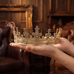 Puede incluir: Una corona de metal dorado con un intrincado diseño de filigrana, motivos florales en relieve a lo largo de la base y elementos verticales puntiagudos y ornamentados que se extienden hacia arriba desde la banda circular, con un acabado antiguo texturizado.