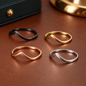 May include: A set of four thin, wave-shaped rings in black, gold, rose gold, and silver tones. Each ring features a smooth, minimalist band with a single V-shaped chevron curve, providing a sleek and uniform appearance across all four metal finishes.
