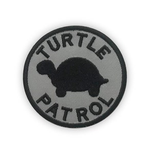 May include: Round embroidered patch with the words "TURTLE PATROL" in black, encircling a black turtle silhouette on a gray background. The patch has a black border and is likely for clothing or accessories.