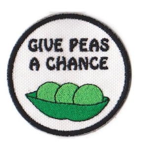 May include: A white round embroidered patch with black stitching. The patch features a green pea pod with three peas inside and the text "Give Peas a Chance".