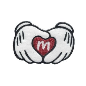 May include: White fabric patch with two hands forming a heart shape. The heart is red with glitter and has a white letter "M" in the center.