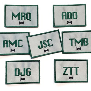 May include: Seven white fabric patches with green borders. Each patch has a different three-letter acronym in green letters, with a black bow tie symbol below each acronym. The acronyms are MRQ, ADD, AMC, JSC, TMB, DJG, and ZTT.