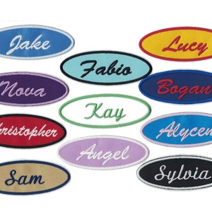 May include: A collection of 11 oval-shaped embroidered patches with various names, including Jake, Lucy, Fabio, Nova, Bogan, Kay, Alycen, Angel, Sam, Sylvia, and Christopher. The patches are in different colors, including blue, yellow, green, purple, red, and black.