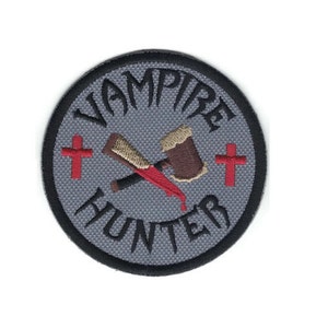 May include: A gray circular embroidered patch with the text "Vampire Hunter" in black lettering around the edge.  Two crossed weapons, a bloody knife and a wooden stake, are in the center of the patch.  Two red crosses are on either side of the weapons.