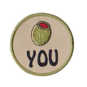 May include: Embroidered patch with a tan background, olive green border, and a green olive with a red center. The word "YOU" is embroidered in black, centered below the olive.