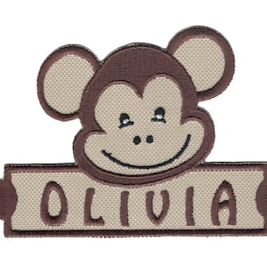 May include: Embroidered patch featuring a brown monkey face with a smile and the name Olivia written below in brown letters on a beige background.