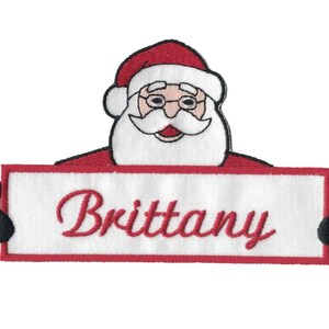 May include: Red and white embroidered Christmas patch with a Santa Claus holding a sign that says "Brittany".