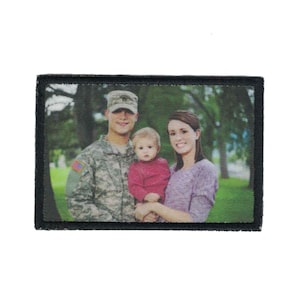 May include: Rectangular patch with a family portrait. The image features a man in a military uniform, a woman in a purple sweater, and a baby in a pink shirt. The patch has a black border and a fabric texture.