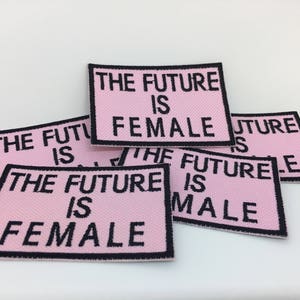 May include: A stack of pink fabric patches with black embroidered text that reads "The Future is Female" and "The Future is Male".