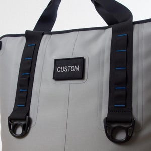 May include: A gray tote bag with black straps and accents. The bag features a black patch with the word "CUSTOM" in white. Blue stitching details the straps. The bag has a sturdy design with metal hardware.