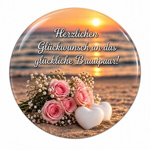 Könnte beinhalten: Ein runder Button mit einem Strandmotiv bei Sonnenuntergang, rosa Rosen, weißem Schleierkraut und zwei weißen herzförmigen Steinen. Der Text lautet: Herzlichen Glückwunsch an das glückliche Brautpaar! Das Design hat eine glänzende Oberfläche.