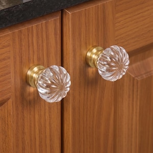 Puede incluir: Dos pomos de armario redondos con cabezas de cristal transparente estriado y textura acanalada. Cada pomo está montado sobre una base metálica circular de tono latón cepillado, fijada a la parte frontal de puertas de madera con veta vertical.