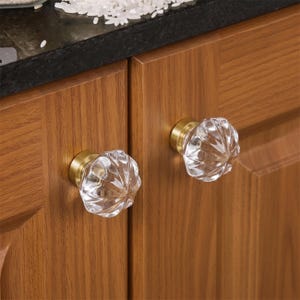 Puede incluir: Dos pomos de armario redondos con tapas de cristal transparente facetado en forma de calabaza festoneada. Cada pomo está montado sobre una base circular de metal en tono latón cepillado, fijado en el frente de dos puertas de madera veteada.