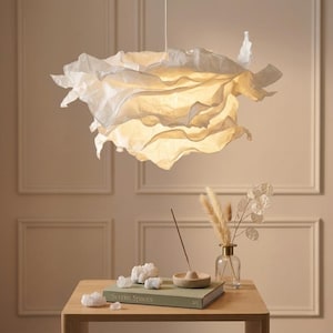 May include: A white pendant light fixture with a layered, ruffled, paper-like shade that emits a warm, soft glow. The shade features organic, undulating edges that create a cloud-like appearance, suspended from a thin cord from the ceiling.