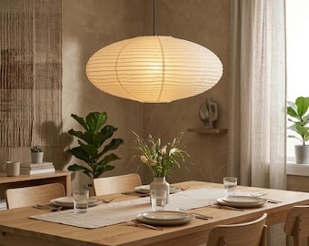 Peut inclure: Une grande suspension ovale dotée d'un abat-jour en papier blanc cassé nervuré, suspendue au plafond. La lampe présente une texture horizontale en couches et diffuse une lueur douce et chaude, centrée au-dessus d'une table à manger en bois.