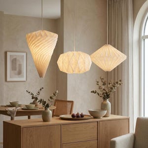 Op de afbeelding: Drie gebroken witte hanglampen met geometrische, geplooide papierachtige texturen hangen aan het plafond. De linkerlamp is kegelvormig, de middelste lamp is bolvormig met een gefacetteerd oppervlak en de rechterlamp is ruitvormig met scherpe randen.