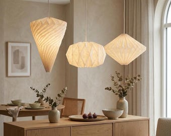 Peut inclure: Trois suspensions blanc cassé avec des textures géométriques plissées semblables à du papier pendent du plafond. La lampe de gauche est en forme de cône, celle du centre est sphérique avec une surface facettée, et celle de droite est en losange.