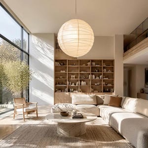 Puede incluir: Una gran lámpara colgante esférica de papel cuelga sobre una mesa de centro redonda de mármol en una sala de estar. La habitación cuenta con un sofá seccional beige, un sillón de madera con detalles de rejilla, una estantería alta y una alfombra.