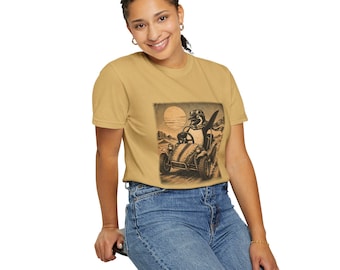 May include: A mustard yellow short-sleeved t-shirt featuring a dark brown graphic print of a penguin wearing goggles and a scarf while driving a small open-top vehicle across a desert landscape with a large sun in the background.