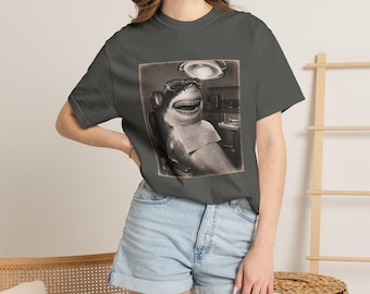 May include: A charcoal gray short-sleeve t-shirt featuring a graphic print of a shark wearing glasses and braces while sitting in a dentist chair. The shark has a wide grin, and a dental bib is draped over its chest under a bright overhead light.