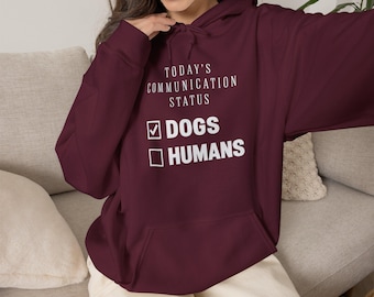 May include: A maroon long-sleeve hooded sweatshirt featuring a front kangaroo pocket and white text that reads: TODAY'S COMMUNICATION STATUS, with a checked box next to DOGS and an unchecked box next to HUMANS, printed in a clean sans-serif font.