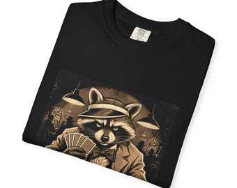 May include: A black t-shirt featuring a graphic print of a raccoon wearing a visor, suit jacket, and bow tie while holding a hand of playing cards. The scene is set in a dimly lit casino environment with slot machines and overhead pendant lights.