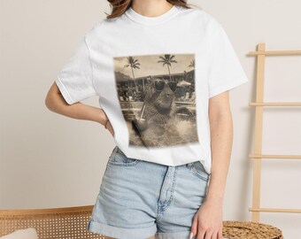 May include: A white short-sleeved t-shirt featuring a sepia-toned graphic print of a capybara wearing sunglasses and holding a drink with a straw, set against a background of palm trees and a building, paired with light blue denim shorts.