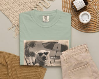 May include: A sage green crewneck t-shirt featuring a graphic print of a polar bear wearing sunglasses, sitting in a beach chair under an umbrella with a palm tree in the background. The shirt has a Comfort Colors brand label at the inner neckline.