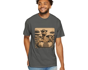 May include: A dark gray t-shirt featuring a graphic illustration of a camel wearing sunglasses, floating in a pool on an inflatable ring while holding a tropical drink. The background shows lounge chairs and umbrellas in a sepia-toned style.