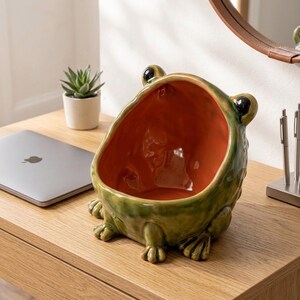 May include: A ceramic frog-shaped bowl with a glossy green exterior and a smooth orange interior. The frog features two prominent black eyes on top and four webbed feet at the base, creating a wide, open-mouthed basin for storage or display.