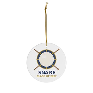 May include: A white circular ceramic ornament featuring a graphic of a blue and gold snare drum with crossed drumsticks. Below the drum, the text reads SNARE in blue and CLASS OF 2027 in gold. The ornament hangs from a thin gold string.