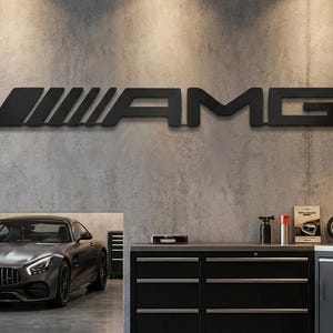 May include: A large, black, wall-mounted AMG logo sign featuring the iconic slanted bars and bold lettering. The sign is displayed on a textured gray concrete wall above a modern garage workspace that includes black cabinets and a gray sports car.