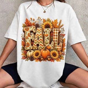 Peut inclure: Un t-shirt blanc à manches courtes avec une grande impression graphique du mot FALL en lettres capitales. Les lettres sont décorées de motifs floraux, chevrons et carreaux, entourées de tournesols, citrouilles, pommes de pin et feuilles.