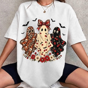 May include: A white short-sleeve t-shirt featuring a graphic print of three ghosts. The left ghost has a leopard print with skulls, the center ghost wears a leopard print bow, and the right ghost is black with red cherries and skulls.