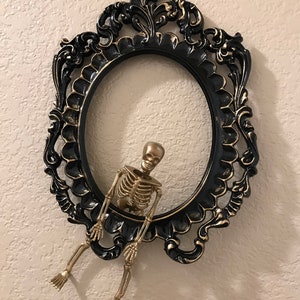 May include: A gold skeleton figurine hanging from a black and gold ornate picture frame.