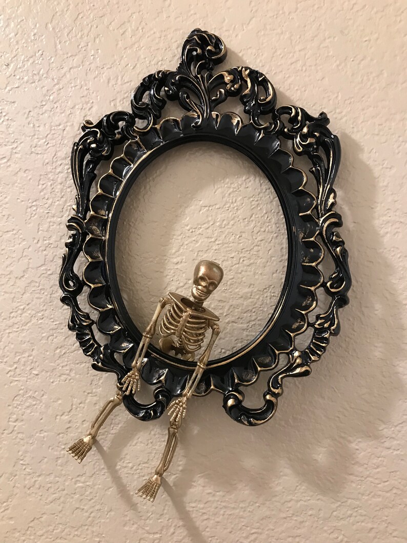 May include: A gold skeleton figurine hanging from a black and gold ornate picture frame.