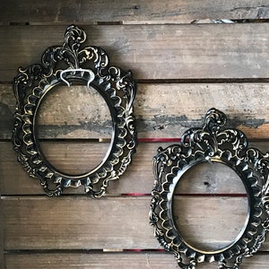 May include: Two ornate black oval picture frames with gold accents hanging on a wooden wall.