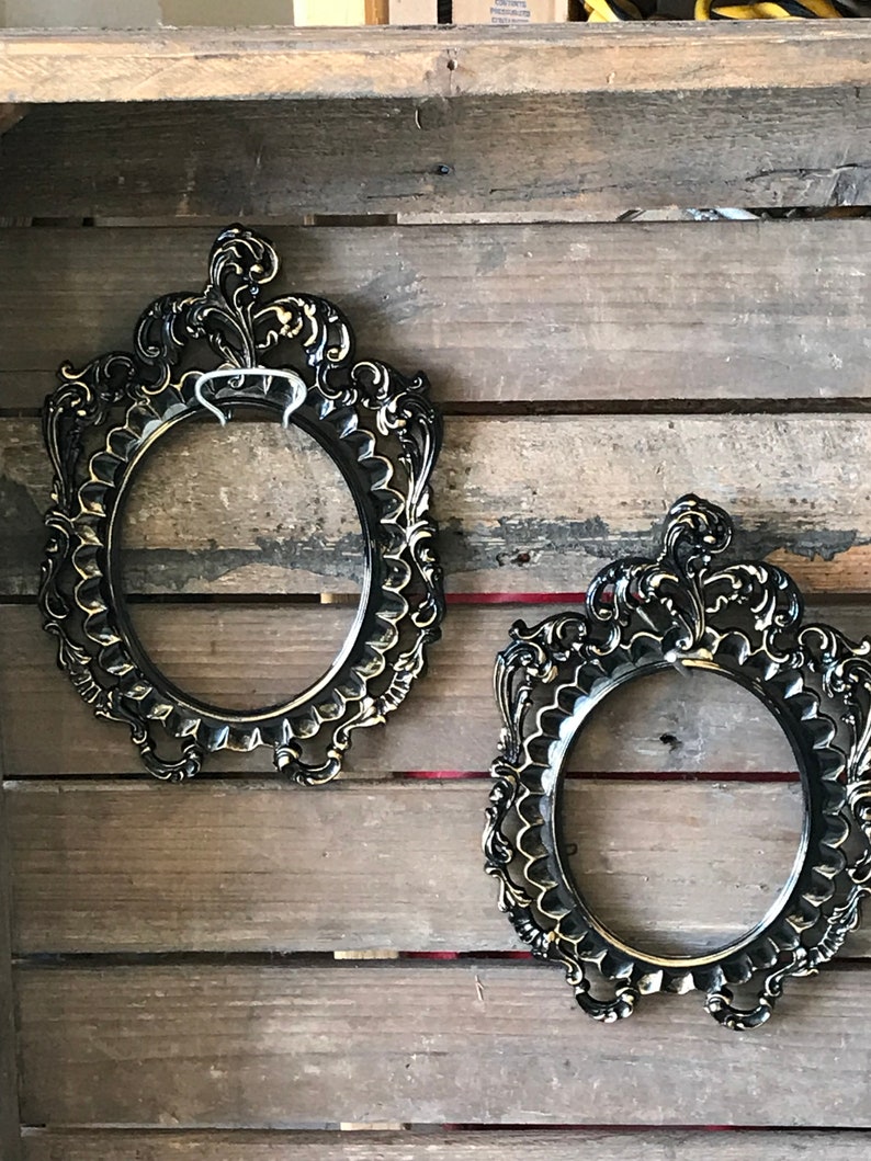 May include: Two ornate black oval picture frames with gold accents hanging on a wooden wall.