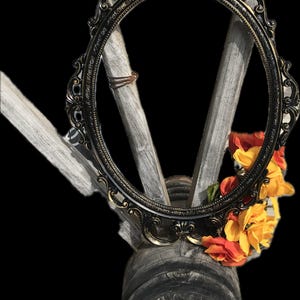 Puede incluir: Marco ovalado ornamentado en negro y dorado con detalles decorativos. El marco se encuentra sobre un fondo de madera envejecida y adornado con flores naranjas y amarillas. El dise&ntilde;o sugiere un tema vintage o r&uacute;stico.