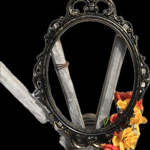 Puede incluir: Marco de fotos ovalado y ornamentado con acabado bronce oscuro. El marco est&aacute; decorado con intrincados adornos y se apoya sobre una base de madera r&uacute;stica. Flores naranjas y amarillas adornan la base, a&ntilde;adiendo color.