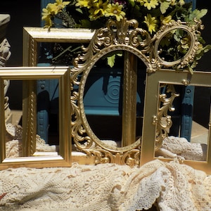 May include: An assortment of gold-colored picture frames in various shapes and sizes. The frames include a square, a rectangle, an oval, and a small round frame. The frames are displayed on a cream-colored lace cloth.