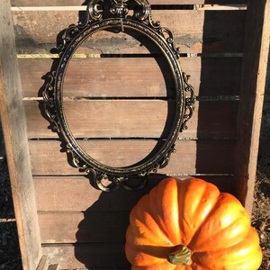 Puede incluir: Un marco ovalado negro y dorado con detalles ornamentales descansa sobre un fondo de madera. Una calabaza naranja brillante se encuentra debajo del marco, sugiriendo un tema oto&ntilde;al. El marco est&aacute; vac&iacute;o, listo para una foto o una obra de arte.