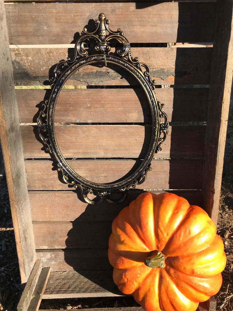 Puede incluir: Un marco ovalado negro y dorado con detalles ornamentales descansa sobre un fondo de madera. Una calabaza naranja brillante se encuentra debajo del marco, sugiriendo un tema oto&ntilde;al. El marco est&aacute; vac&iacute;o, listo para una foto o una obra de arte.