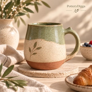 May include: A ceramic mug featuring a speckled cream body with a sage green glazed upper section and a terracotta base. The mug displays a small hand-painted leaf sprig design and includes the text PotteryDigga in the upper right corner.